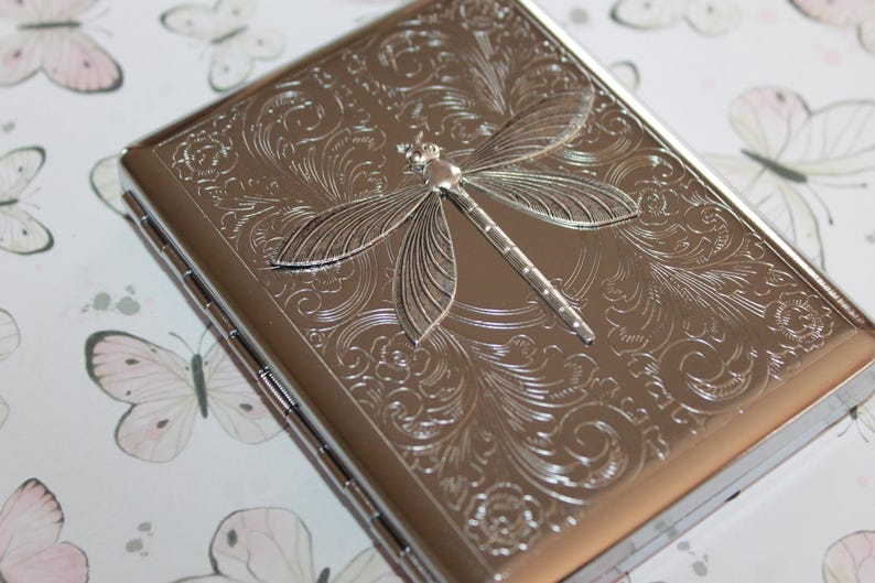 May include: A silver-colored rectangular case with a detailed dragonfly design on the top. The dragonfly is in the center, with intricate floral patterns surrounding it. The case has a shiny, metallic finish.