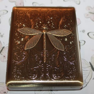 May include: A bronze-coloured metal case with a dragonfly motif. The case features an embossed floral pattern and a dragonfly with outstretched wings. The case is likely for storing small items, such as cigarettes or cards.