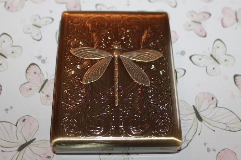 May include: A bronze-coloured metal case with a dragonfly motif. The case features an embossed floral pattern and a dragonfly with outstretched wings. The case is likely for storing small items, such as cigarettes or cards.