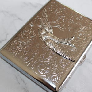 Puede incluir: Cajetilla de cigarrillos de metal plateado con un dise&ntilde;o en relieve detallado de un p&aacute;jaro y flores. La caja tiene forma rectangular con bordes redondeados y un cierre de broche. El p&aacute;jaro est&aacute; en vuelo, con las alas extendidas. El fondo es un patr&oacute;n floral arremolinado.