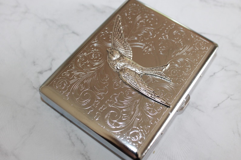 Puede incluir: Cajetilla de cigarrillos de metal plateado con un dise&ntilde;o en relieve detallado de un p&aacute;jaro y flores. La caja tiene forma rectangular con bordes redondeados y un cierre de broche. El p&aacute;jaro est&aacute; en vuelo, con las alas extendidas. El fondo es un patr&oacute;n floral arremolinado.