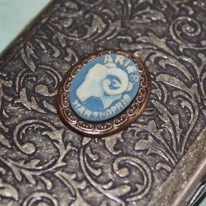 Puede incluir: Broche ovalada con camafeo de Aries, con fondo azul claro y dise&ntilde;o de carnero blanco. La broche est&aacute; engarzada en un marco color cobre y descansa sobre una superficie plateada, ornamentada y estampada. El texto "ARIES MAR 21-APR 19" es visible.