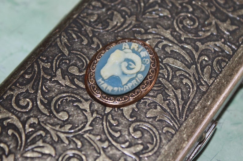 Puede incluir: Broche ovalada con camafeo de Aries, con fondo azul claro y dise&ntilde;o de carnero blanco. La broche est&aacute; engarzada en un marco color cobre y descansa sobre una superficie plateada, ornamentada y estampada. El texto "ARIES MAR 21-APR 19" es visible.