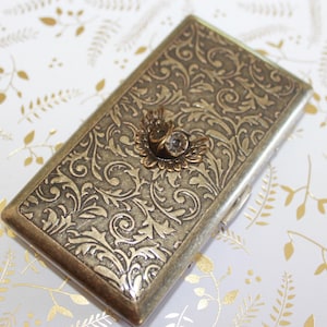 May include: A rectangular metal case with an antique brass finish, featuring an intricate embossed floral scrollwork pattern across the surface and a small, centered owl emblem with outstretched wings and a clear crystal accent on the lid.