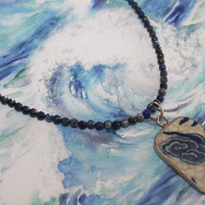 May include: A necklace featuring a string of small, faceted dark blue lapis lazuli beads and a rectangular pendant. The pendant is made of white ceramic with a crackled glaze finish, decorated with a blue painted cloud pattern and a metal border.