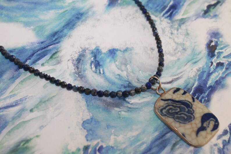 May include: A necklace featuring a string of small, faceted dark blue lapis lazuli beads and a rectangular pendant. The pendant is made of white ceramic with a crackled glaze finish, decorated with a blue painted cloud pattern and a metal border.