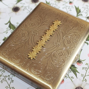 May include: A rectangular brass-toned metal case featuring an intricate engraved floral scrollwork pattern across the surface. A vertical decorative strip of small, connected gold-toned flower shapes is centered on the top of the case.