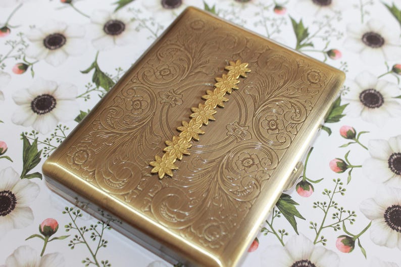 May include: A rectangular brass-toned metal case featuring an intricate engraved floral scrollwork pattern across the surface. A vertical decorative strip of small, connected gold-toned flower shapes is centered on the top of the case.