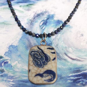 May include: A necklace featuring a string of small, faceted dark blue beads and a rectangular pendant. The pendant is made of white ceramic with a blue floral pattern and a crackled glaze finish, encased in a simple silver-toned metal frame.
