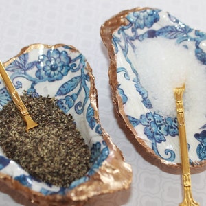 May include: Two white ceramic oyster shells with gold accents, each containing a small gold spoon and a different seasoning. One shell holds salt and the other holds pepper.