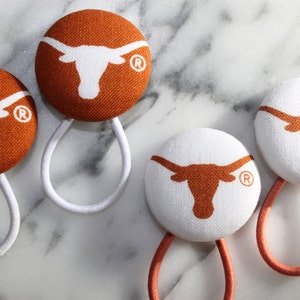 May include: Four hair ties with orange and white fabric covered buttons featuring a white Longhorn steer logo with a registered trademark symbol. The hair ties are white, orange, and pink.