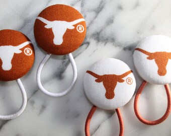 May include: Four hair ties with orange and white fabric covered buttons featuring a white Longhorn steer logo with a registered trademark symbol. The hair ties are white, orange, and pink.
