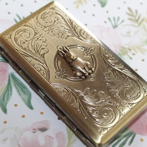Puede incluir: Un estuche rectangular de metal en tono lat&oacute;n con una figura de conejo en relieve en el centro, rodeada de intrincados patrones de volutas. El estuche tiene un lado con bisagras y un acabado met&aacute;lico con un dise&ntilde;o de superficie texturizado.