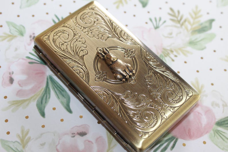 Puede incluir: Un estuche rectangular de metal en tono lat&oacute;n con una figura de conejo en relieve en el centro, rodeada de intrincados patrones de volutas. El estuche tiene un lado con bisagras y un acabado met&aacute;lico con un dise&ntilde;o de superficie texturizado.