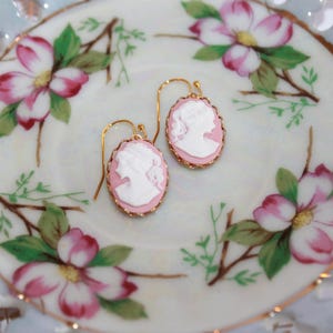 May include: A pair of pink cameo earrings with gold-tone settings. The earrings are displayed on a white porcelain plate with a floral design.