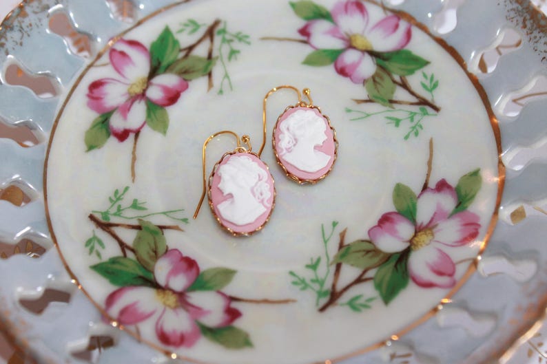May include: A pair of pink cameo earrings with gold-tone settings. The earrings are displayed on a white porcelain plate with a floral design.
