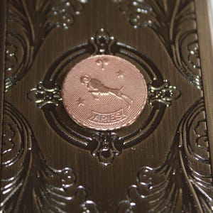 May include: A bronze-colored metal box with ornate floral and scrollwork. A copper-colored circular emblem in the center features the Aries zodiac symbol and the word "ARIES".