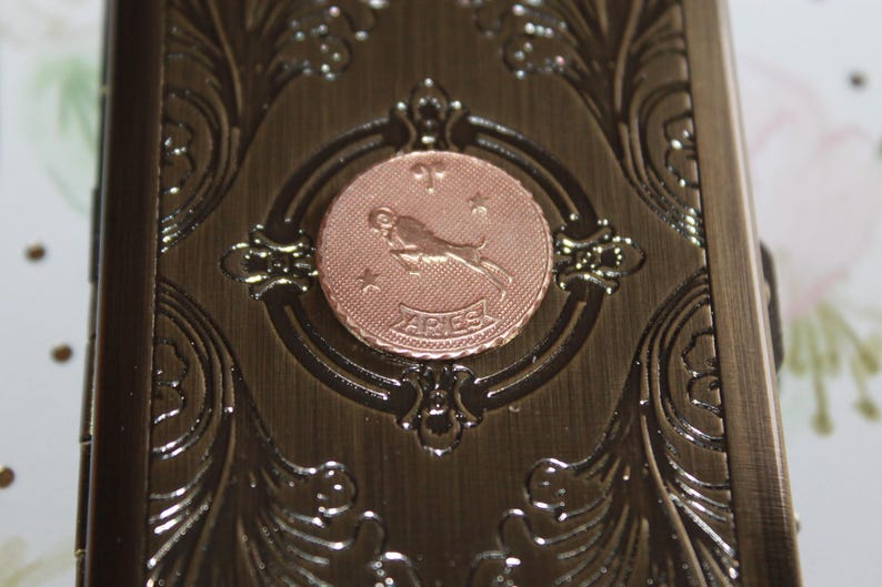May include: A bronze-colored metal box with ornate floral and scrollwork. A copper-colored circular emblem in the center features the Aries zodiac symbol and the word "ARIES".