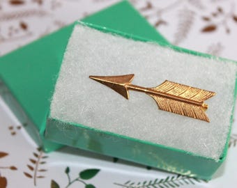 May include: A gold-toned metal arrow pin featuring a pointed tip and textured fletching, resting inside a small, rectangular, bright green cardboard box lined with a thick layer of white foam padding for secure storage and display.