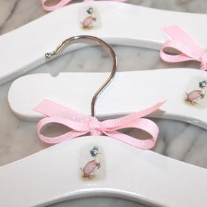May include: White wooden hangers with a pink ribbon and a printed image of a duck wearing a dress.