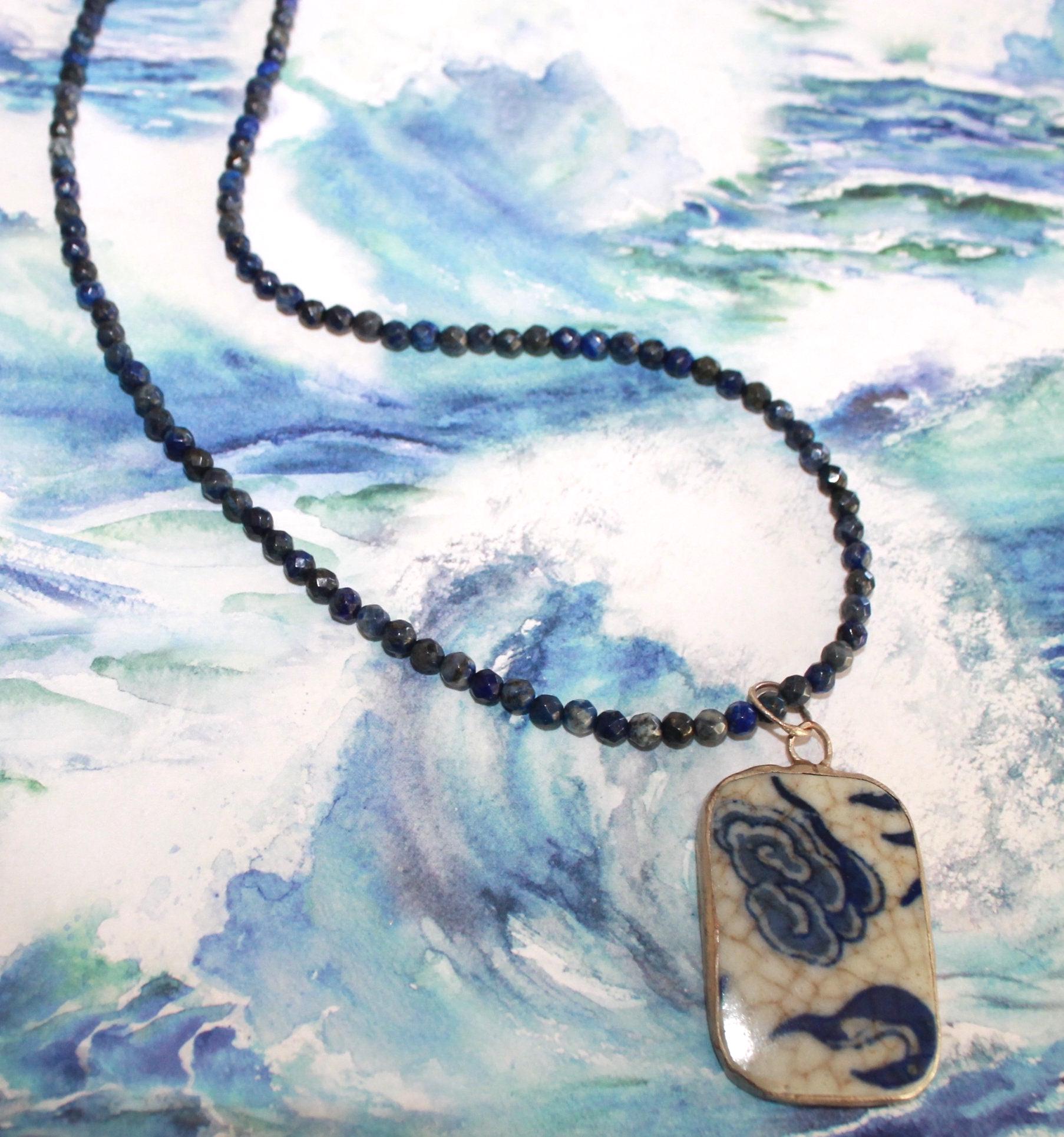 May include: A necklace featuring a strand of small, faceted, dark blue lapis lazuli beads and a rectangular pendant. The pendant has a cream-colored base with a blue, hand-painted floral pattern and a visible crackled glaze finish on the surface.