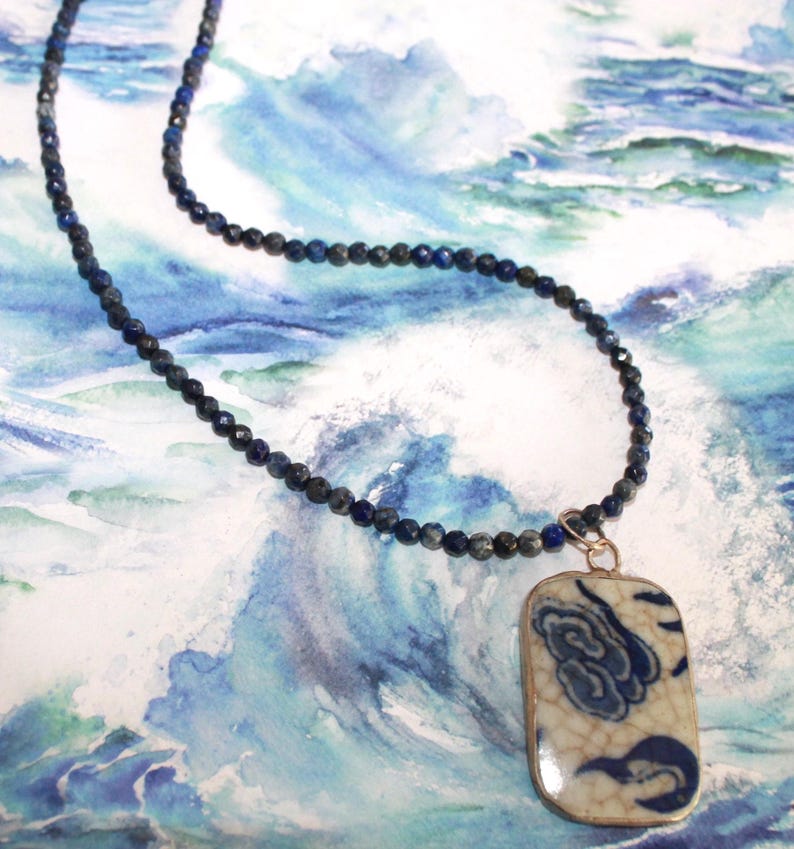 May include: A necklace featuring a strand of small, faceted, dark blue lapis lazuli beads and a rectangular pendant. The pendant has a cream-colored base with a blue, hand-painted floral pattern and a visible crackled glaze finish on the surface.