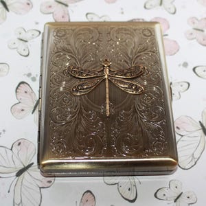 Puede incluir: Una pitillera rectangular de metal con acabado en bronce cepillado. La superficie frontal presenta una lib&eacute;lula en relieve centrada sobre intrincados patrones de volutas y flores. El estuche tiene un lado con bisagras y un marco met&aacute;lico.