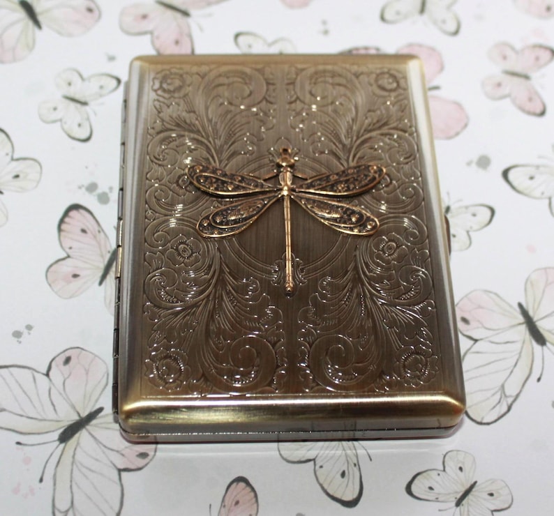 Puede incluir: Una pitillera rectangular de metal con acabado en bronce cepillado. La superficie frontal presenta una lib&eacute;lula en relieve centrada sobre intrincados patrones de volutas y flores. El estuche tiene un lado con bisagras y un marco met&aacute;lico.