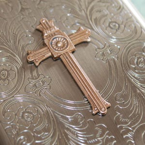 Puede incluir: Un estuche met&aacute;lico rectangular con acabado en bronce cepillado y un intrincado patr&oacute;n floral grabado. Un emblema de cruz en tono cobre est&aacute; centrado en la tapa, mostrando las letras INRI en la parte superior y un detalle circular.
