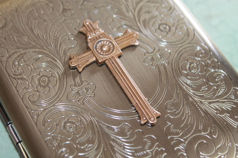 Puede incluir: Un estuche met&aacute;lico rectangular con acabado en bronce cepillado y un intrincado patr&oacute;n floral grabado. Un emblema de cruz en tono cobre est&aacute; centrado en la tapa, mostrando las letras INRI en la parte superior y un detalle circular.