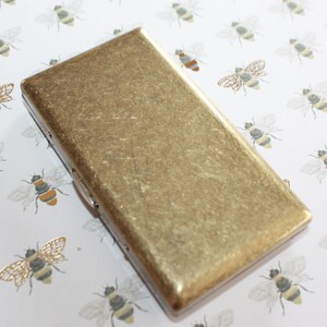 May include: A gold metal cigarette case with a textured surface. The case is on a white background with a pattern of bees.
