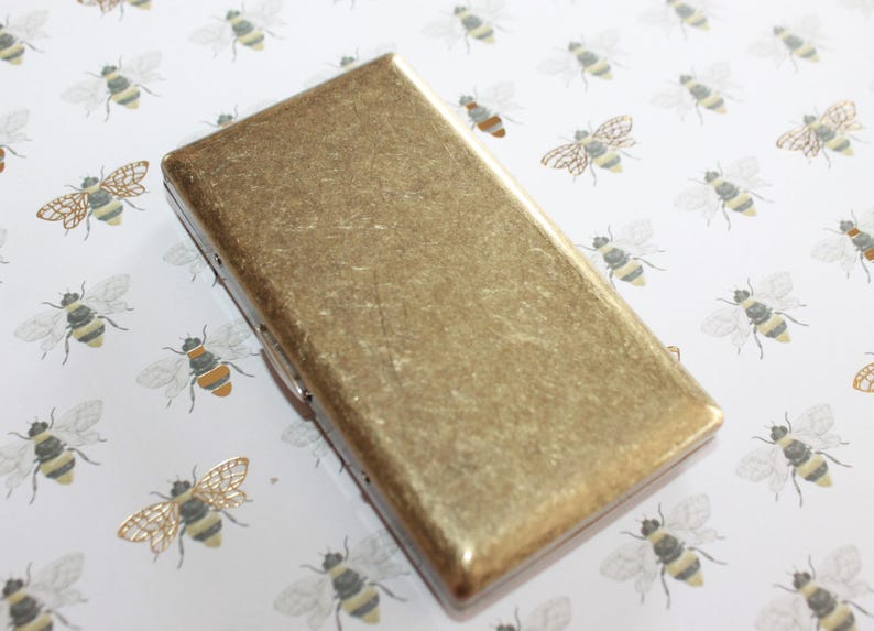 May include: A gold metal cigarette case with a textured surface. The case is on a white background with a pattern of bees.