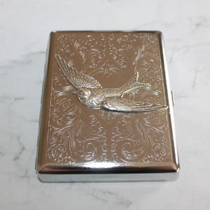 Puede incluir: Un estuche de cigarrillos de metal plateado con un dise&ntilde;o en relieve. El estuche presenta un relieve detallado de un p&aacute;jaro en vuelo, sobre un fondo de intrincados patrones florales. El estuche es rectangular con bordes redondeados.