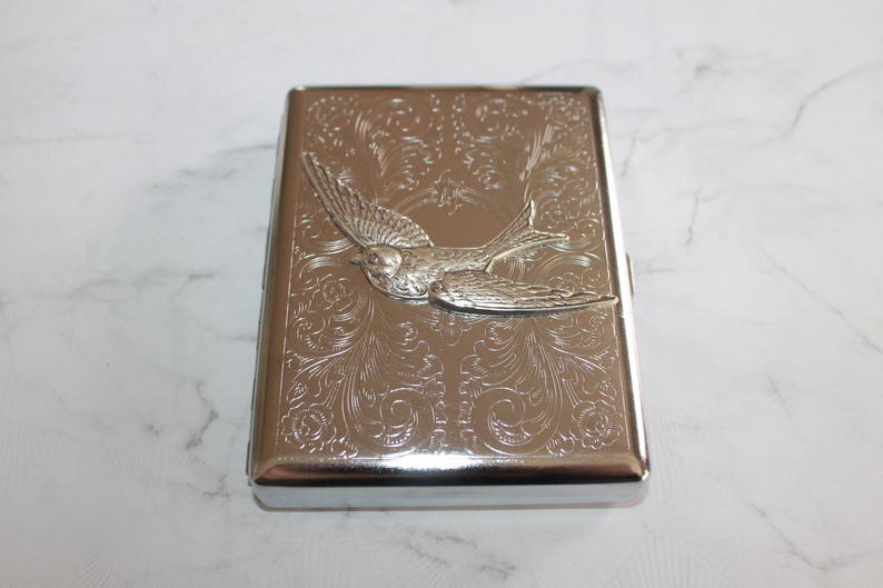 Puede incluir: Un estuche de cigarrillos de metal plateado con un dise&ntilde;o en relieve. El estuche presenta un relieve detallado de un p&aacute;jaro en vuelo, sobre un fondo de intrincados patrones florales. El estuche es rectangular con bordes redondeados.