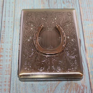 May include: A rectangular metal cigarette case with an embossed floral design and a horseshoe-shaped clasp. The case has a bronze finish and is set against a blue and white wooden background.