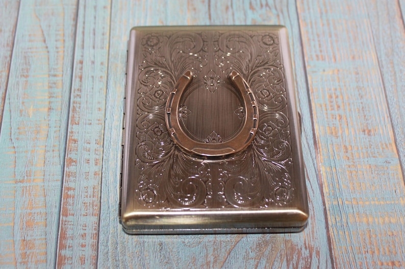 May include: A rectangular metal cigarette case with an embossed floral design and a horseshoe-shaped clasp. The case has a bronze finish and is set against a blue and white wooden background.