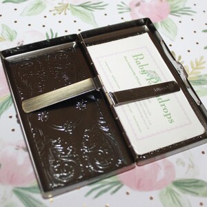 May include: Open, dark brown metal business card holder with an embossed floral design. The holder is open, revealing white cards with green text that reads "Baby Props". The background is a floral pattern.