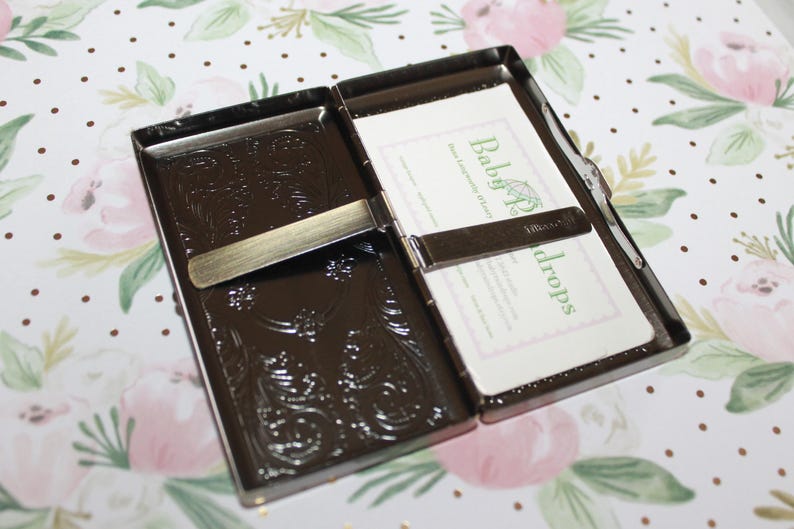 May include: Open, dark brown metal business card holder with an embossed floral design. The holder is open, revealing white cards with green text that reads "Baby Props". The background is a floral pattern.