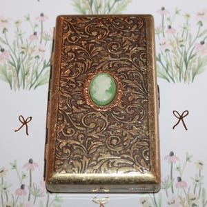 May include: A gold metal cigarette case with an ornate floral design and a green cameo with a white silhouette of a person's profile.