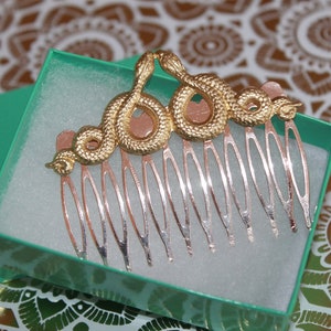 May include: A gold snake comb with a silver comb. The snake is curled up and has a detailed design. The comb is in a green box.