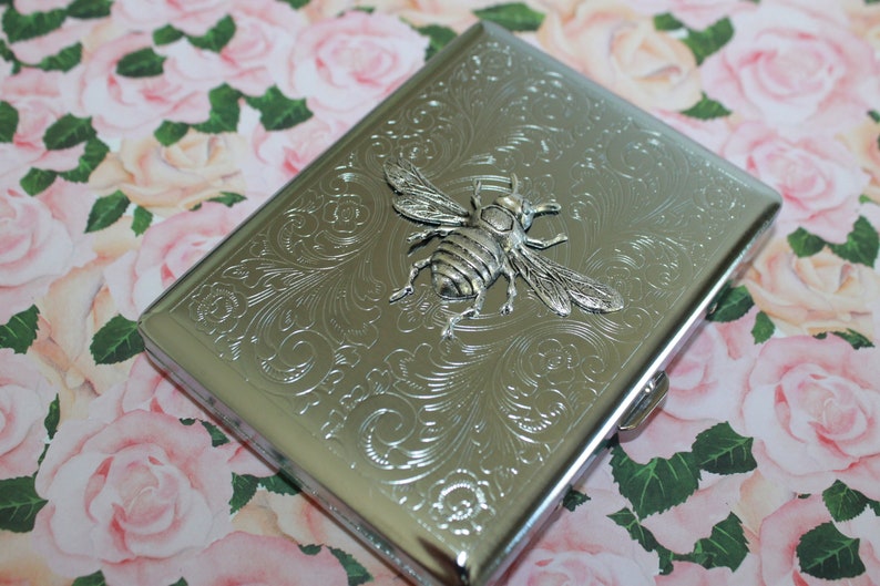 May include: Silver metal cigarette case with an embossed bee design. The case is rectangular with rounded corners and a clasp. The surface is decorated with floral patterns. The bee is in the centre of the case.