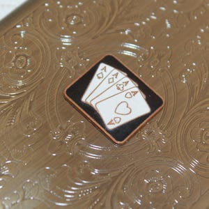 K&ouml;nnte beinhalten: Ein rechteckiges Etui aus goldfarbenem Metall mit einem gepr&auml;gten floralen Rankenmuster auf der Oberfl&auml;che. In der Mitte des Deckels befindet sich eine rautenf&ouml;rmige Einlage mit schwarzem Hintergrund und vier wei&szlig;en Spielkarten-Assen.