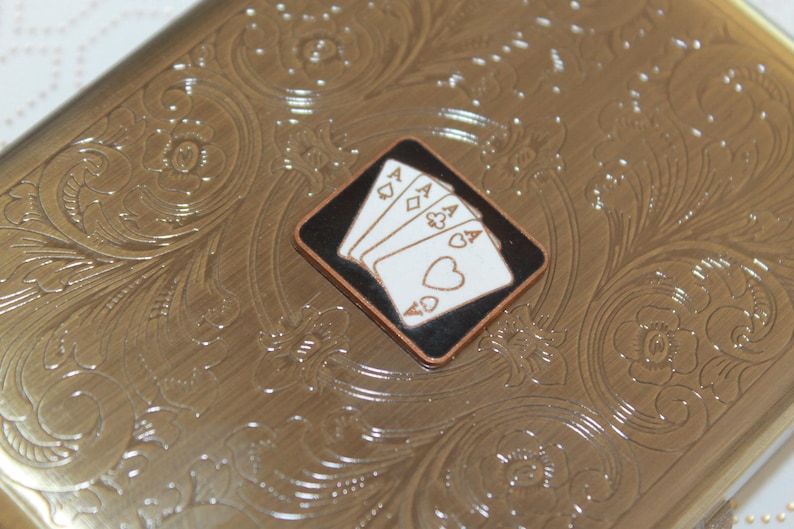 K&ouml;nnte beinhalten: Ein rechteckiges Etui aus goldfarbenem Metall mit einem gepr&auml;gten floralen Rankenmuster auf der Oberfl&auml;che. In der Mitte des Deckels befindet sich eine rautenf&ouml;rmige Einlage mit schwarzem Hintergrund und vier wei&szlig;en Spielkarten-Assen.