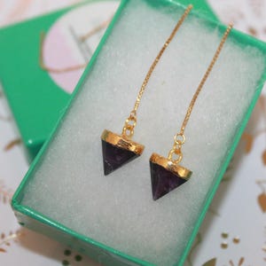 May include: A pair of gold dangle earrings with purple triangle-shaped gemstone pendants. The earrings are displayed in a green gift box.