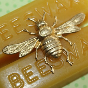 Może przedstawiać: Dwa bloki wosku pszczelego, jeden z wytłoczonym napisem "BEESWAX", a drugi z wytłoczonym napisem "BEESWAX". Na blokach umieszczona jest złota figurka pszczoły.
