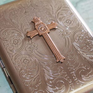 Puede incluir: Un estuche met&aacute;lico rectangular con acabado en bronce cepillado, que presenta un intrincado patr&oacute;n floral en relieve en toda la superficie y un prominente emblema de cruz tridimensional centrado en la tapa frontal del contenedor con bisagras.