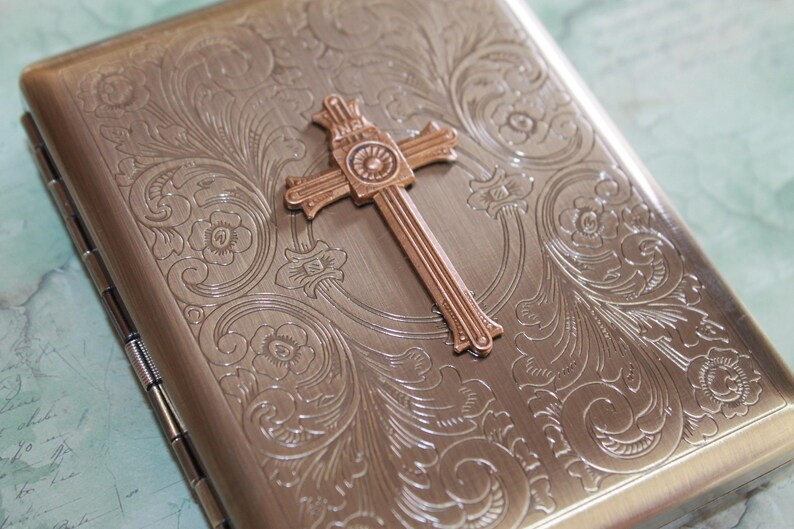 Puede incluir: Un estuche met&aacute;lico rectangular con acabado en bronce cepillado, que presenta un intrincado patr&oacute;n floral en relieve en toda la superficie y un prominente emblema de cruz tridimensional centrado en la tapa frontal del contenedor con bisagras.