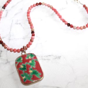 May include: A necklace featuring a strand of small, faceted pink and reddish beads. The pendant is a rectangular metal frame containing a red enamel inlay decorated with a repeating pattern of green leafy vines and small yellow accents.