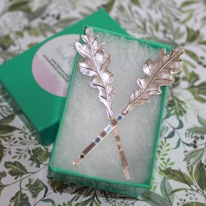 May include: Two silver-colored hair pins shaped like oak leaves are displayed in a green box with a white interior. The pins are crossed, showcasing their intricate leaf details. The box sits on a floral patterned surface.