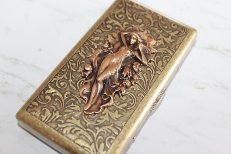 May include: A gold metal cigarette case with an ornate floral design and a copper-colored sculpted figure of a woman.